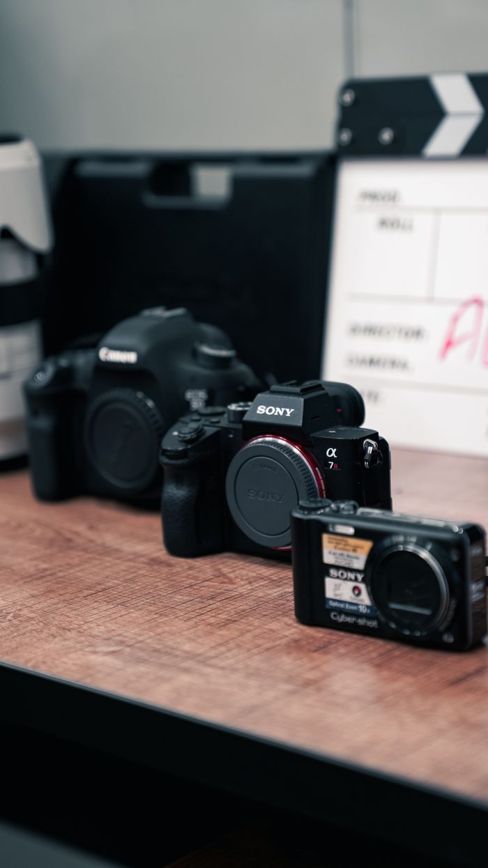 Three cameras of different sizes and brands are showcased on a wooden table in a film set environment.